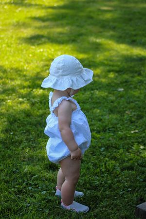 Child in white ruffled outfit taking steps on green grass in summer sunlight.