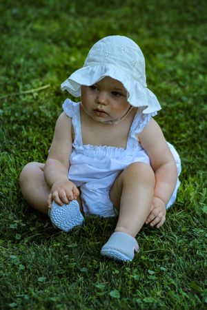 Small child in white summer outfit sitting on green grass.