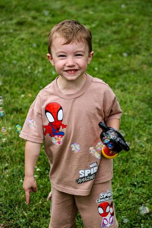 Child wearing tan Spider-Man t-shirt playing in grassy outdoor area.