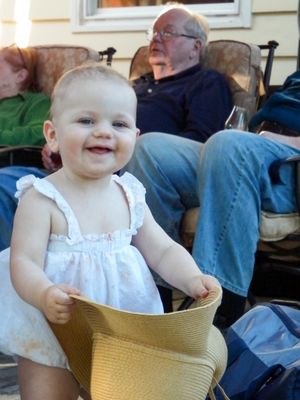 Baby wearing white dress sitting in yellow chair smiling during outdoor gathering.