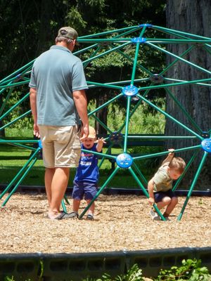 Children playing on blue and green spider web climbing structure at playground.