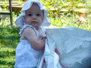 Baby wearing white sunhat and dress sitting outside in summer.