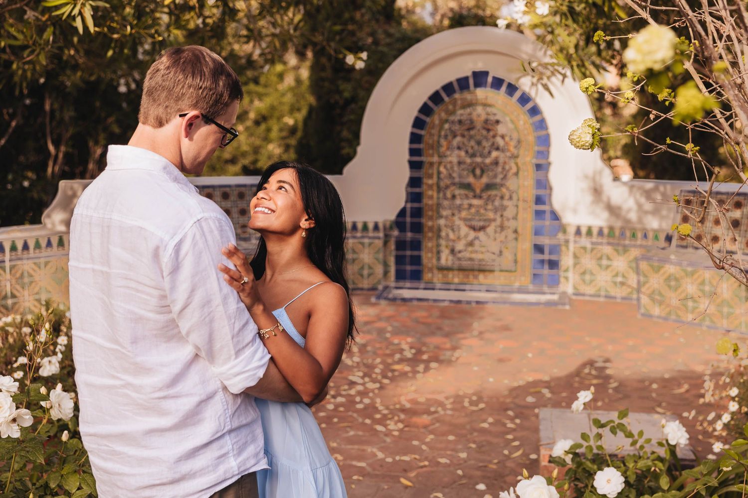 A couple embraces outdoors during golden hour sunlight in a garden setting near flowering orange trees.