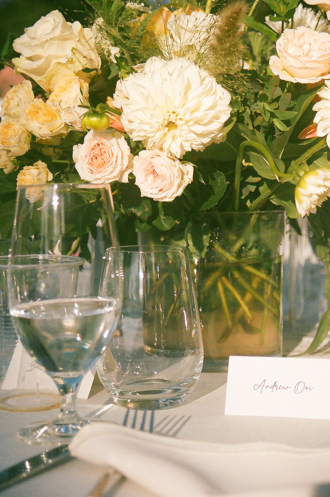 An elegant table setting with peach and cream roses in glass vases, wine glasses, and place cards for a formal event.