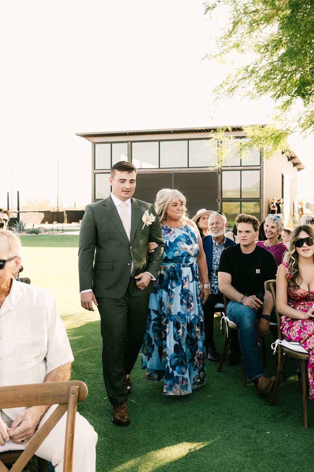 Outdoor wedding reception with guests seated on wooden chairs on manicured lawn.