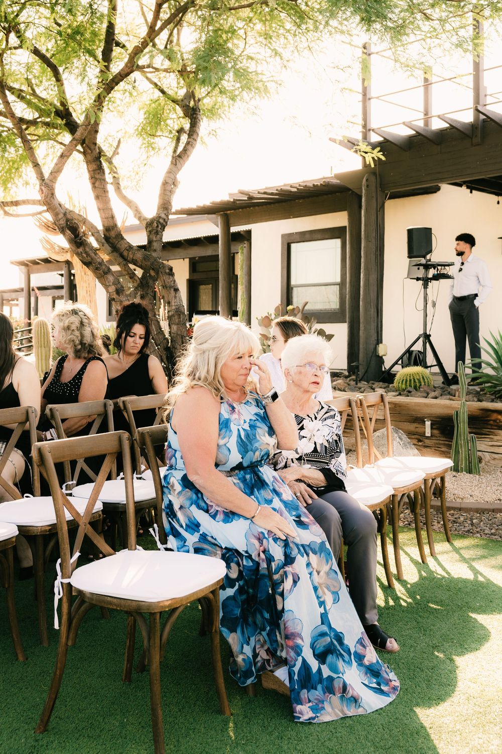 Wedding guests in blue floral dresses seated on wooden chairs at outdoor ceremony.