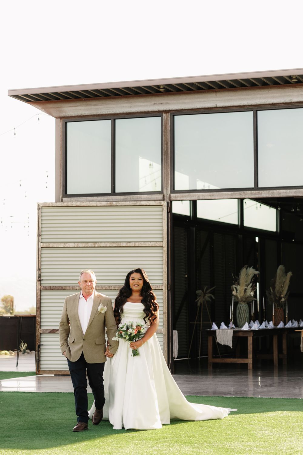 Bride in white dress walks with escort across a green lawn outside a modern building.