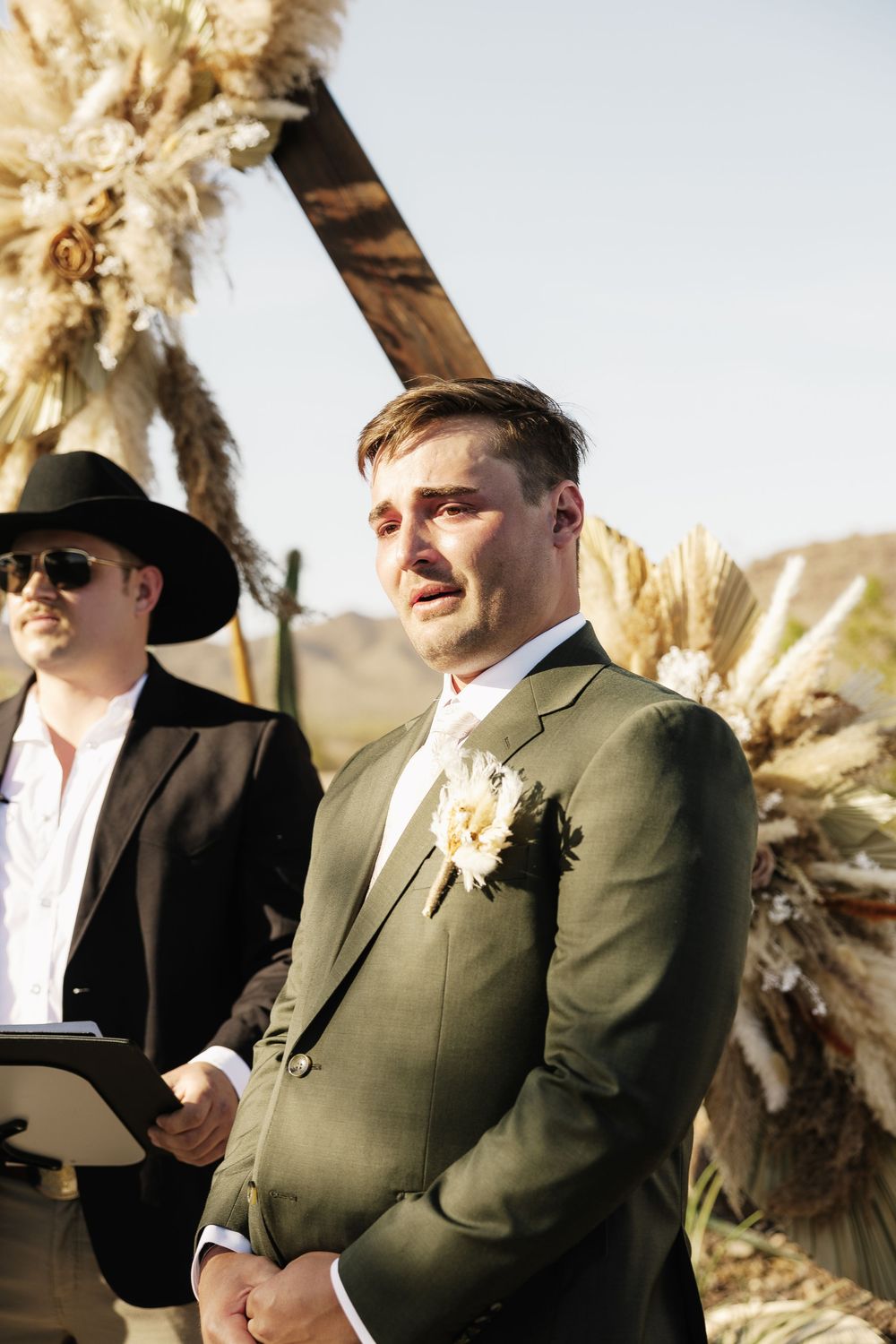 Wedding officiant in green suit and black hat speaks during outdoor desert ceremony.