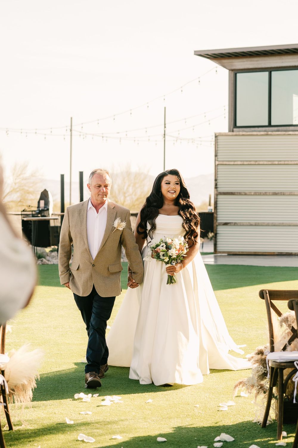 Wedding ceremony processional on green lawn with modern building in background.