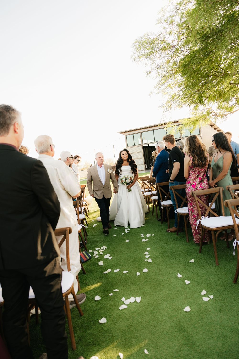 Wedding guests seated on wooden chairs with flower petals scattered on green lawn.
