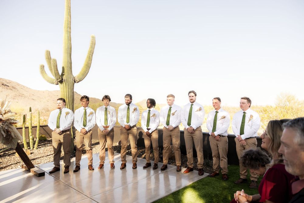 Groomsmen in light colored suits stand in line against a desert landscape with tall saguaro cacti.