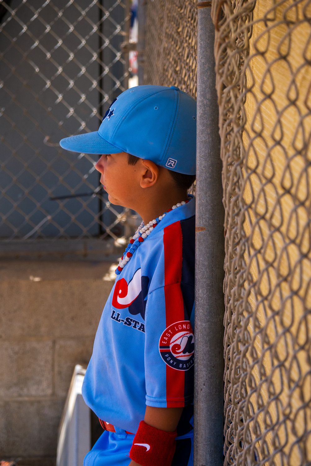 Youth baseball player waits in dugout wearing light blue uniform with red accents.