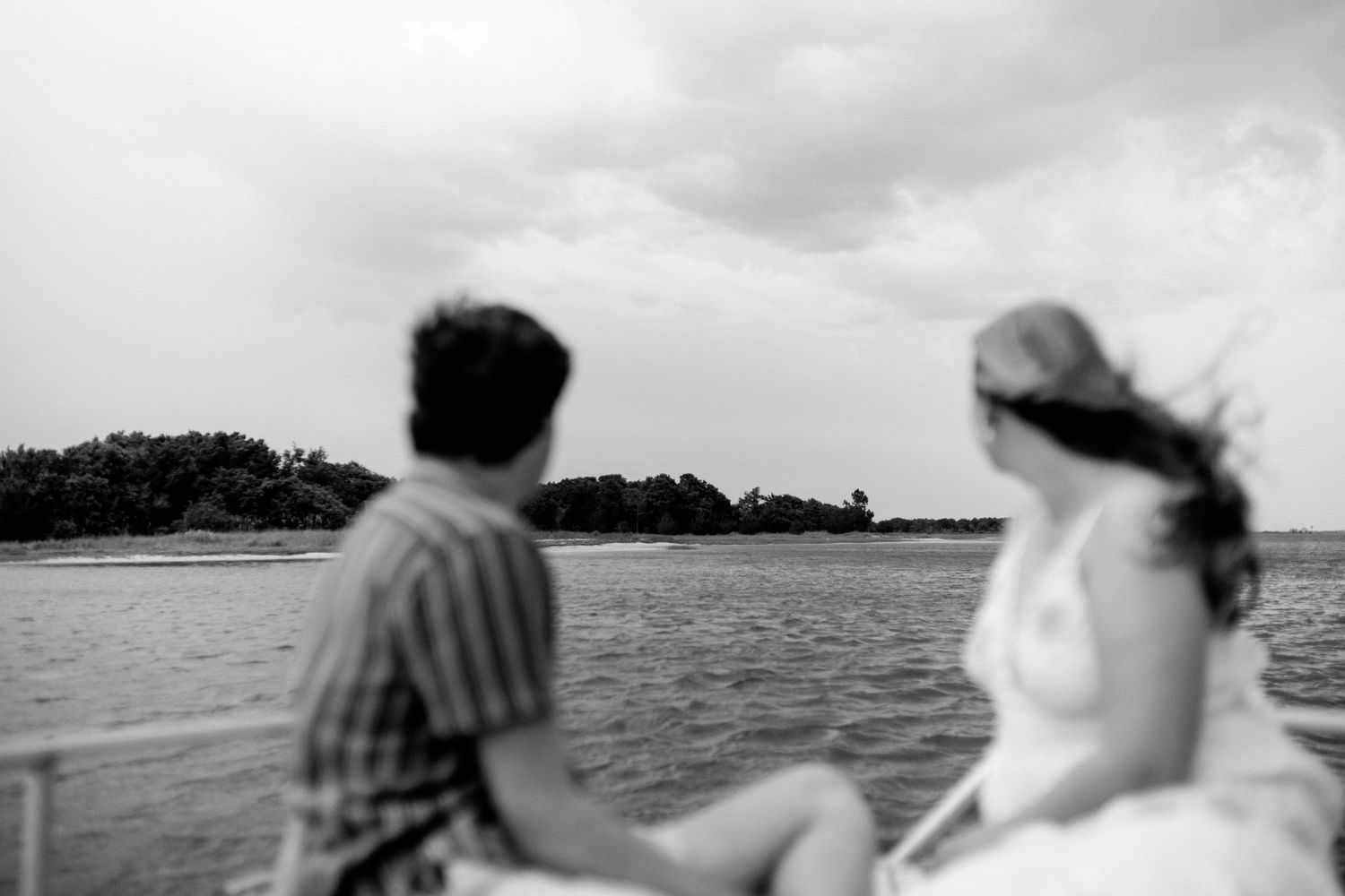 Two people sit together overlooking a serene lake with trees in the background in black and white.