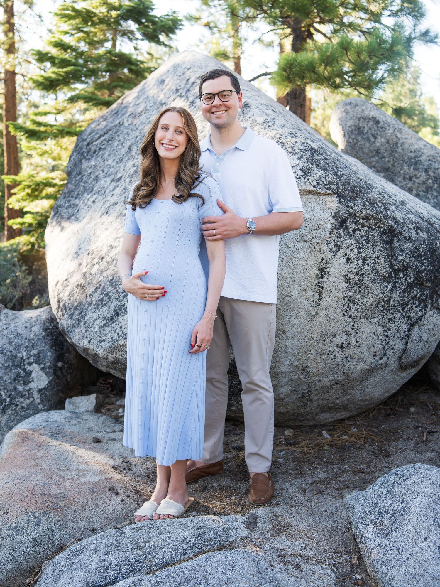 A couple poses together on large granite boulders surrounded by tall pine trees on a sunny day.