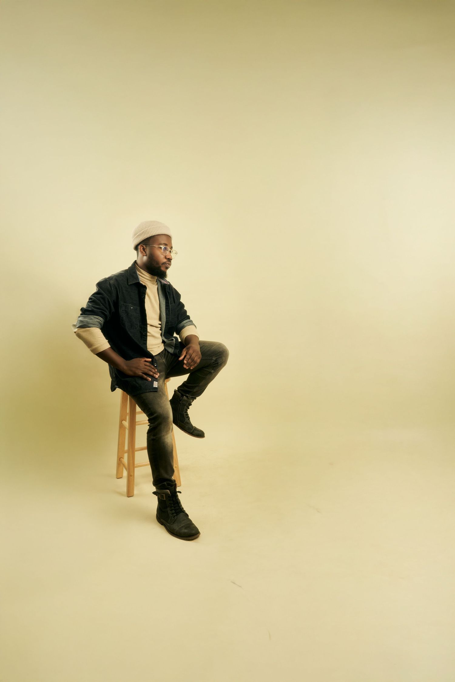 Leonard Okor in black jacket and boots seated on wooden stool against beige backdrop in studio setting.