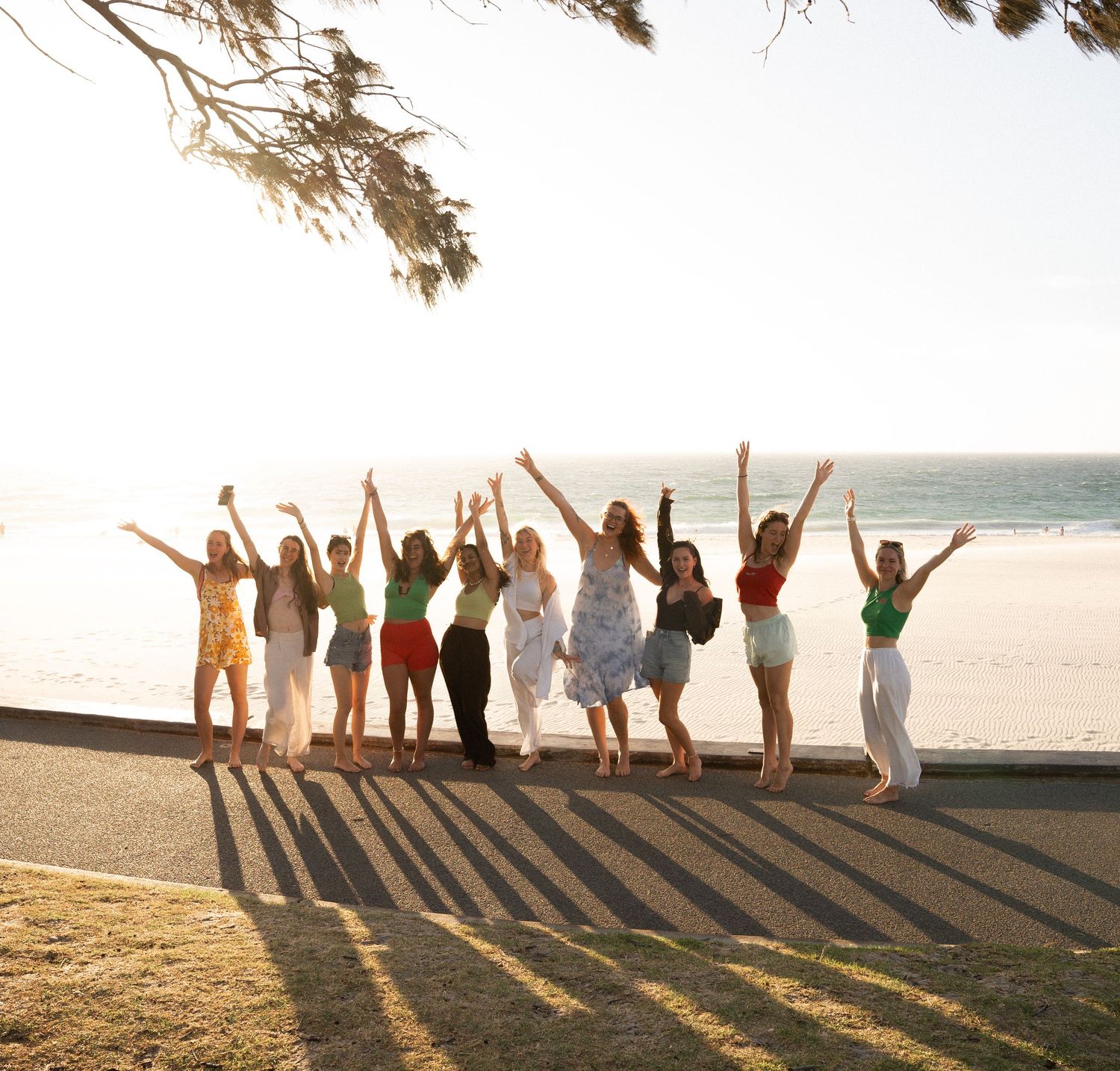 Group of people raising their arms in celebration on a beach at sunset creating long shadows in the sand.