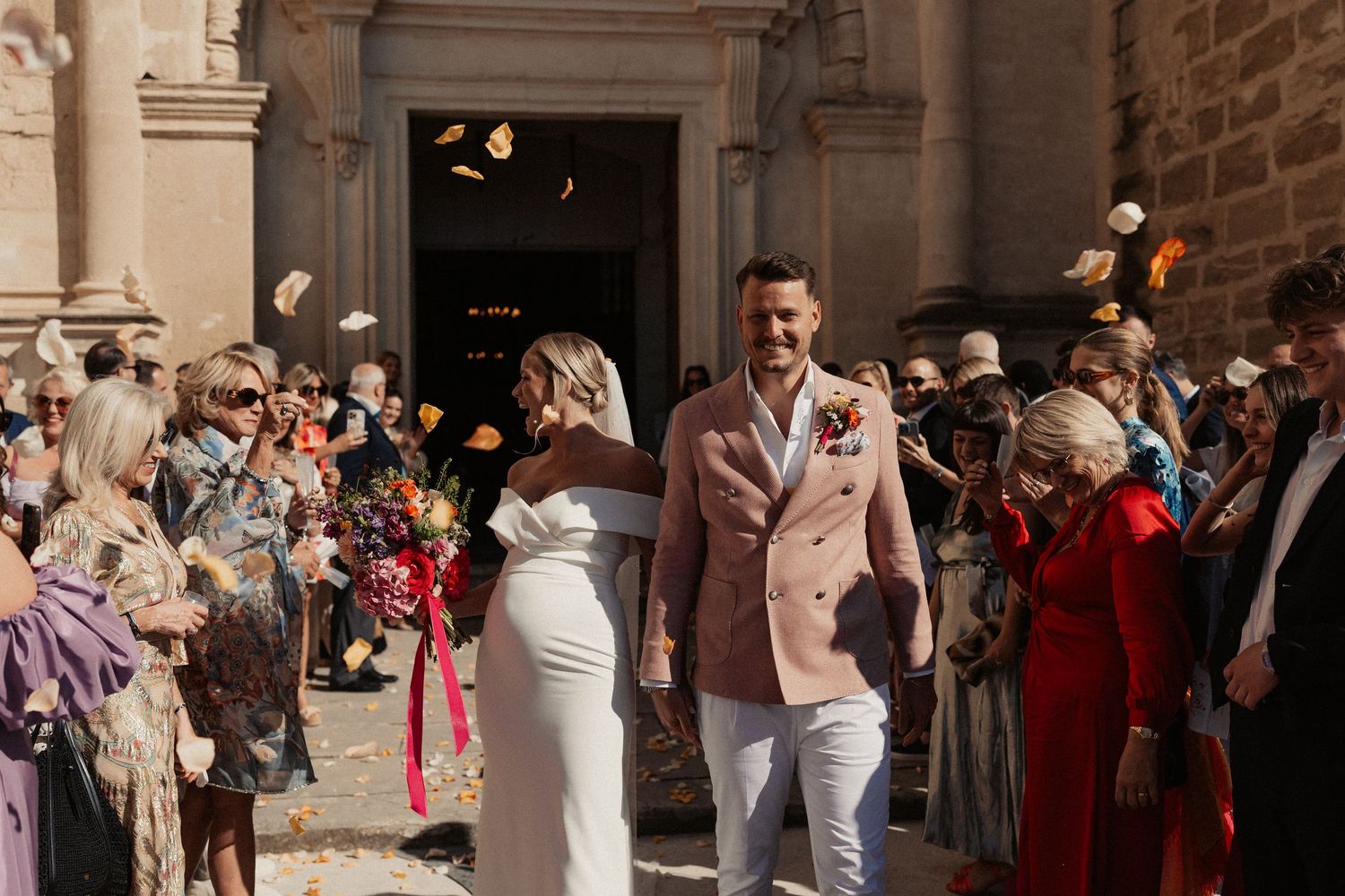 A crowd of wedding guests stands outside a stone church throwing flower petals into the air during a celebration.