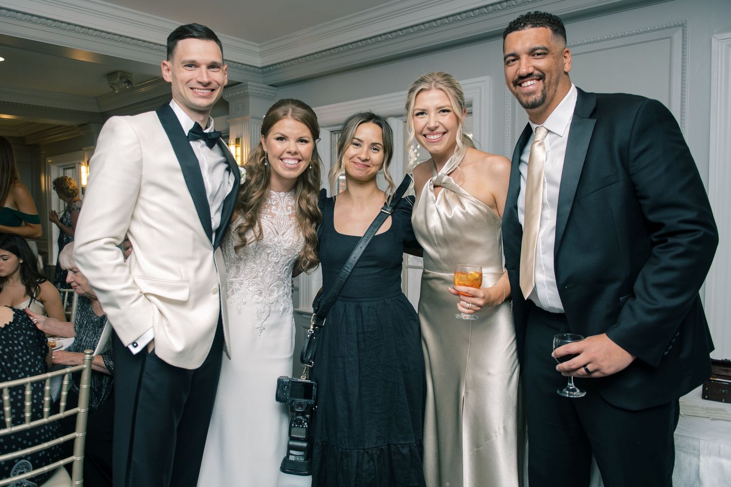A group celebrates together at an elegant wedding reception, with people dressed in formal attire including tuxedos and gowns.
