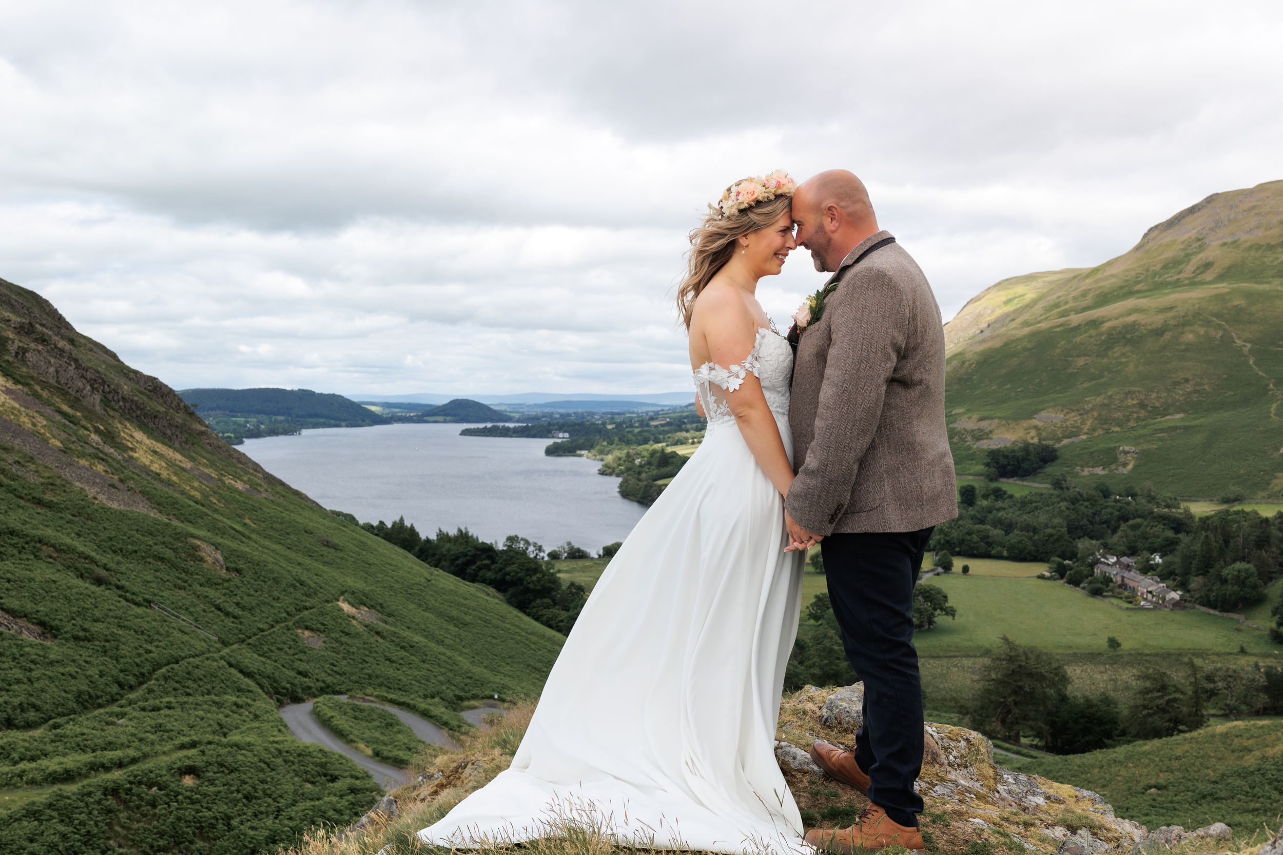 A couple in wedding attire stands on a hilltop overlooking a scenic valley with rolling green hills and distant mountains.