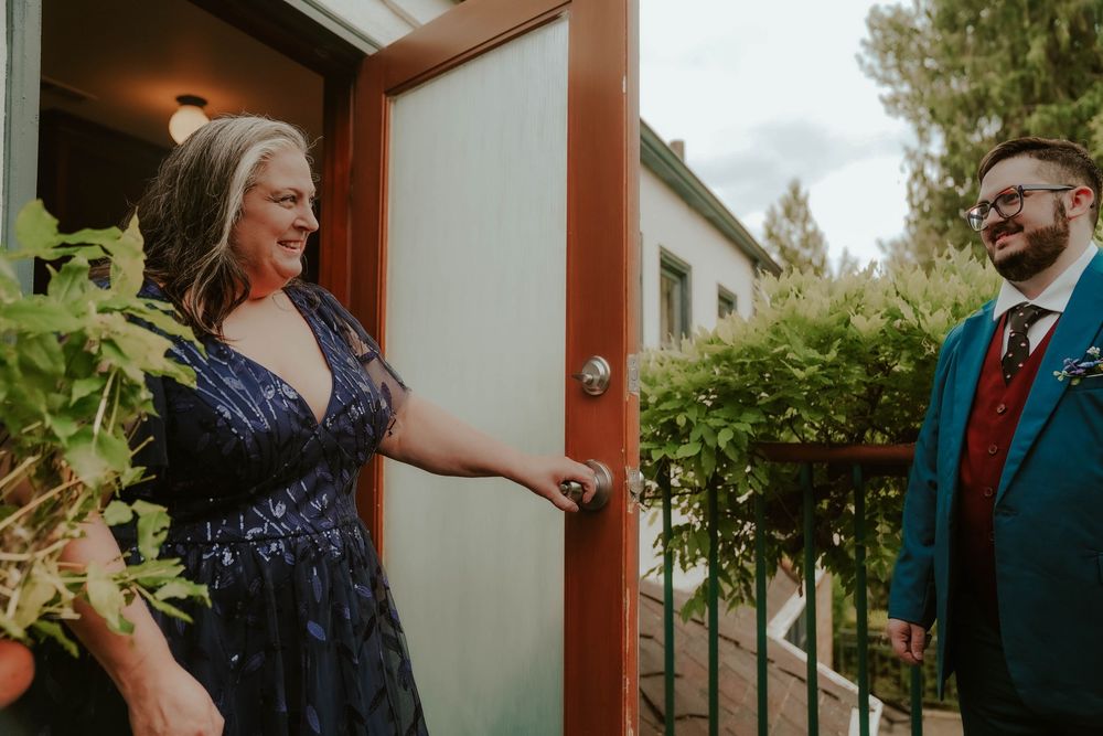A woman in a navy dress opens a door to greet a smiling man in a teal suit at a wedding.
