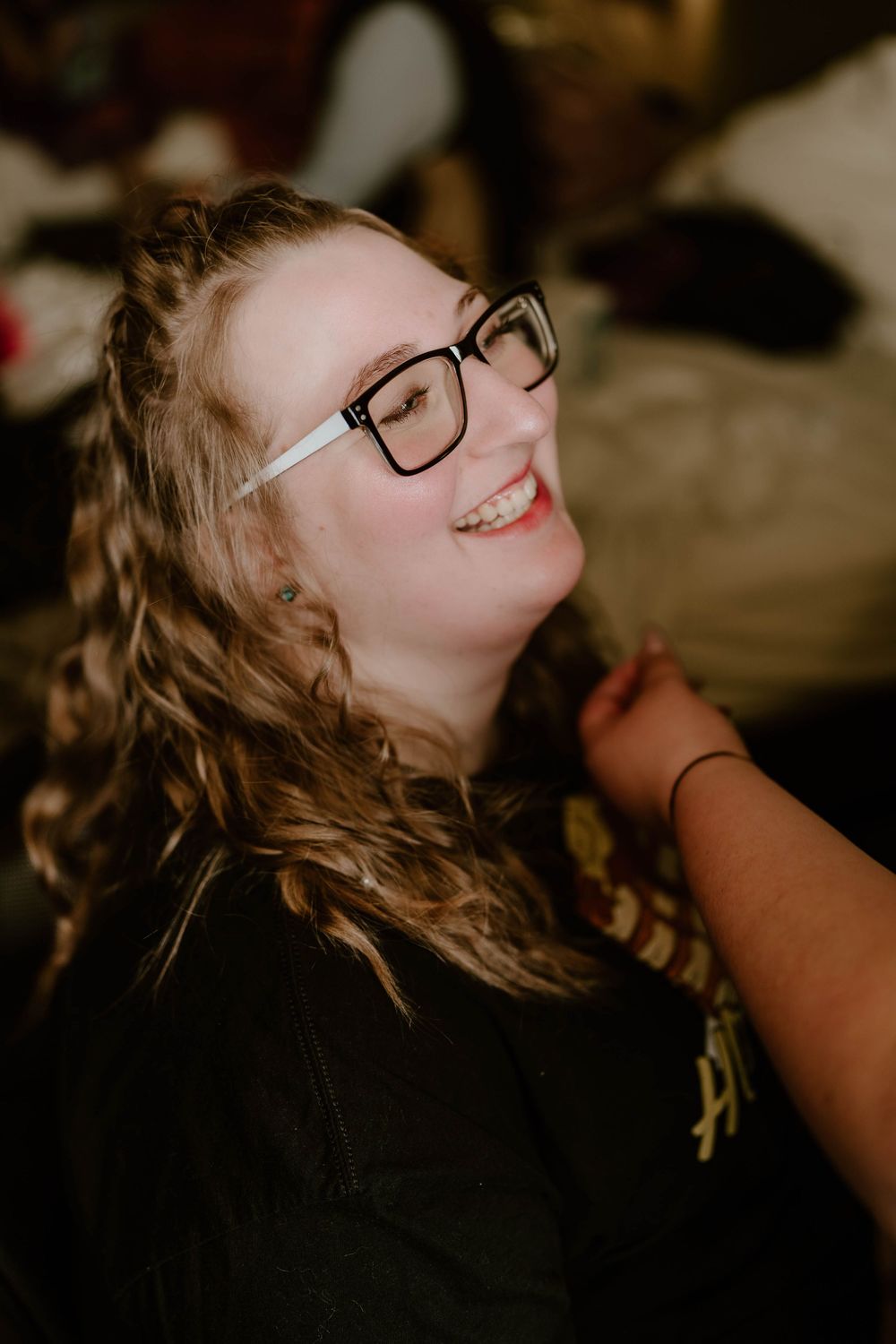 A young woman with curly hair and black glasses smiles brightly while getting ready for a wedding.