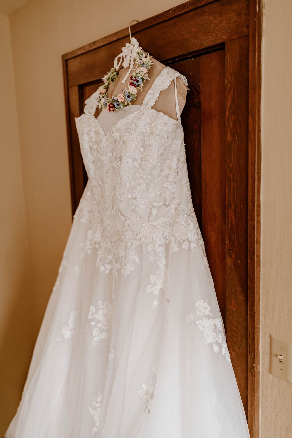 An elegant white lace A-line wedding dress hanging on a decorative floral hanger on a wooden door.