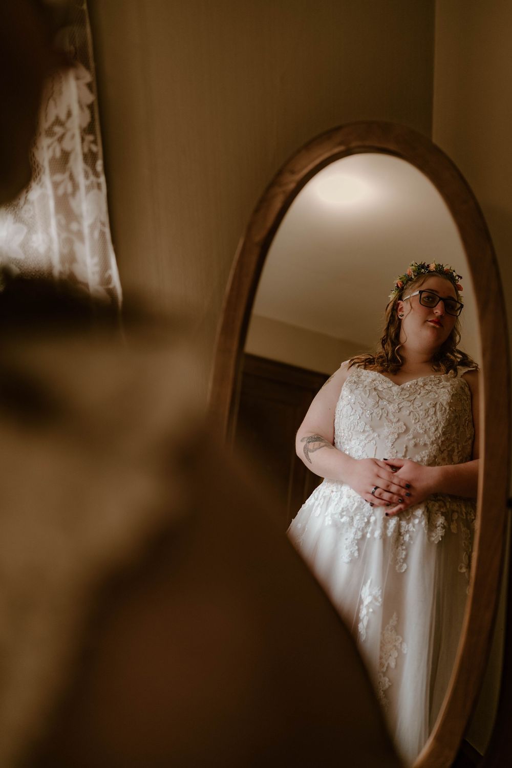 Bride in lace wedding dress and floral crown reflected in vintage oval mirror in warm-lit room.
