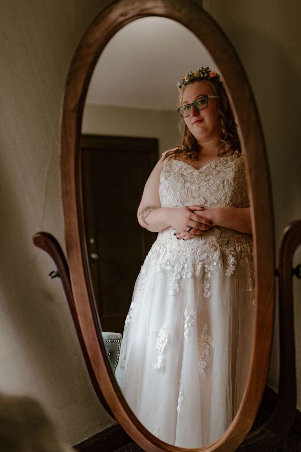 A bride in a white lace gown and floral crown reflects in a vintage oval wooden mirror.