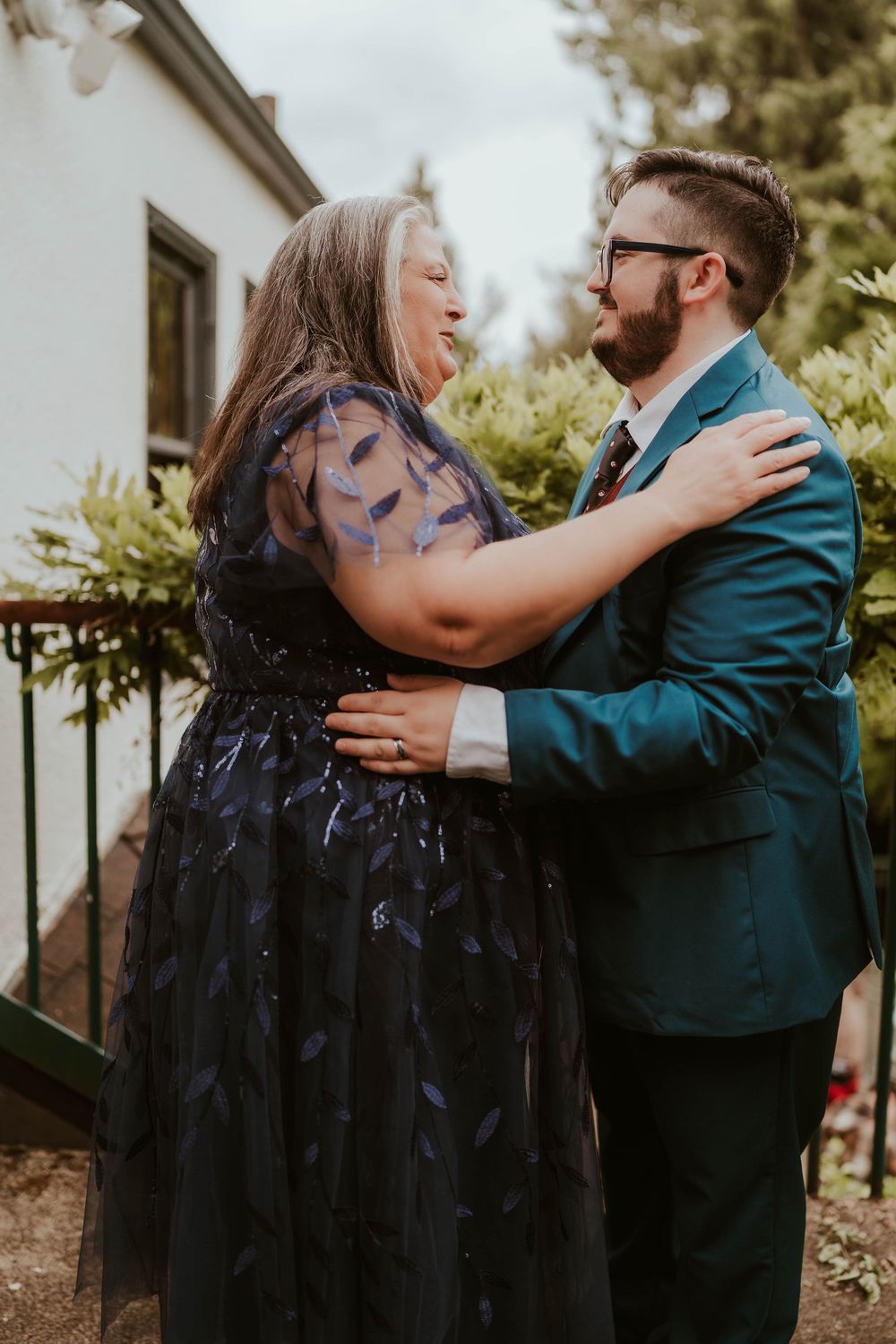 A woman in a navy blue dress and a man in a teal suit share a warm embrace outdoors at a wedding.
