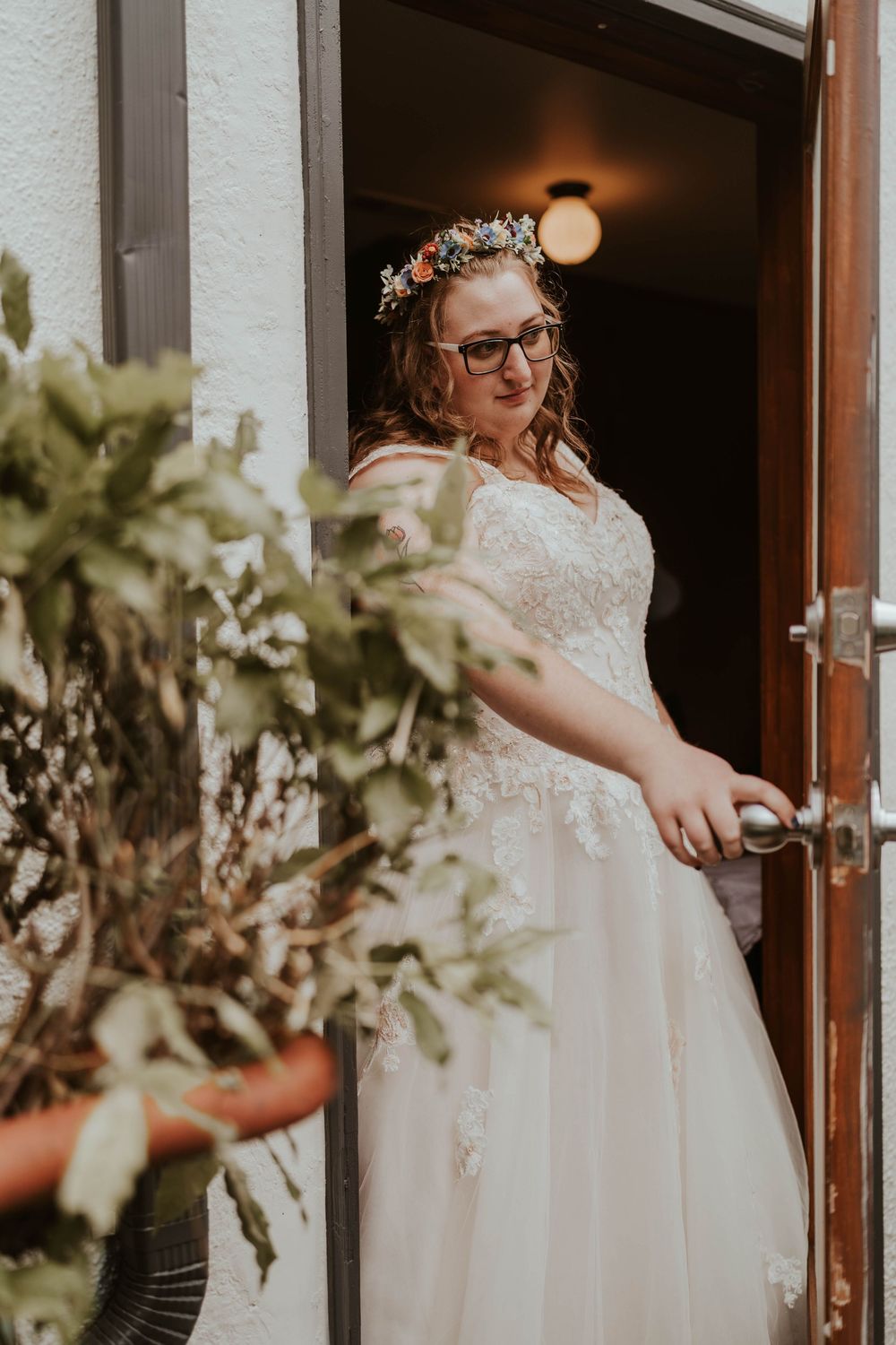 A bride wearing glasses and a floral crown stands in a white lace wedding gown at a doorway.