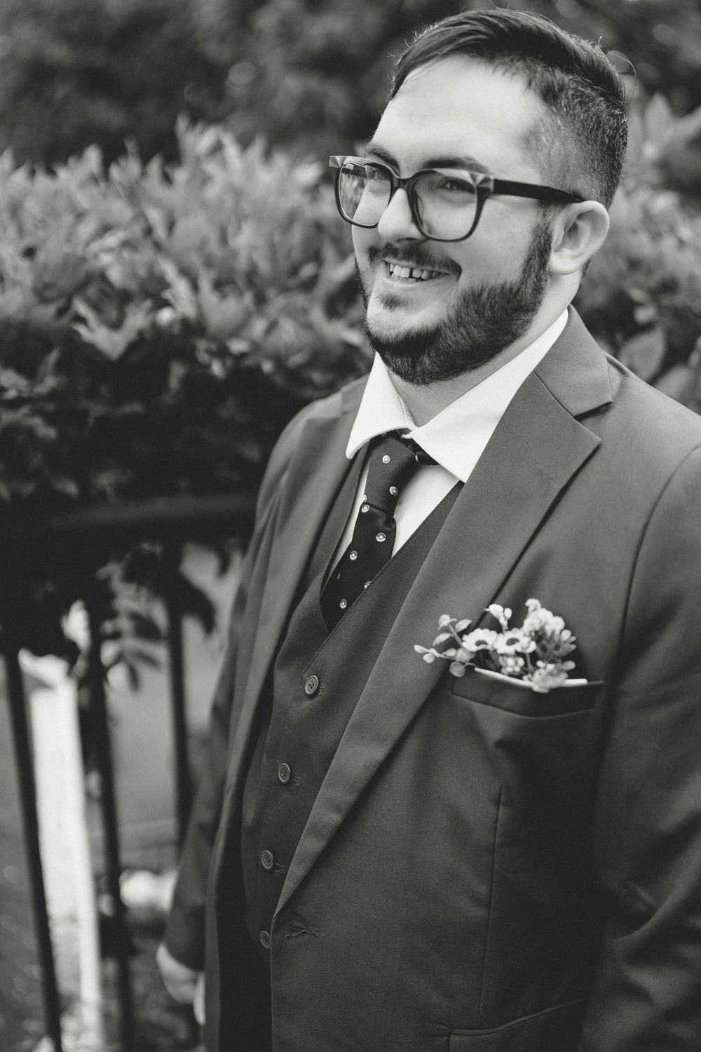 Smiling bearded groom in three-piece suit with floral pocket square and polka dot tie, black and white wedding photo.