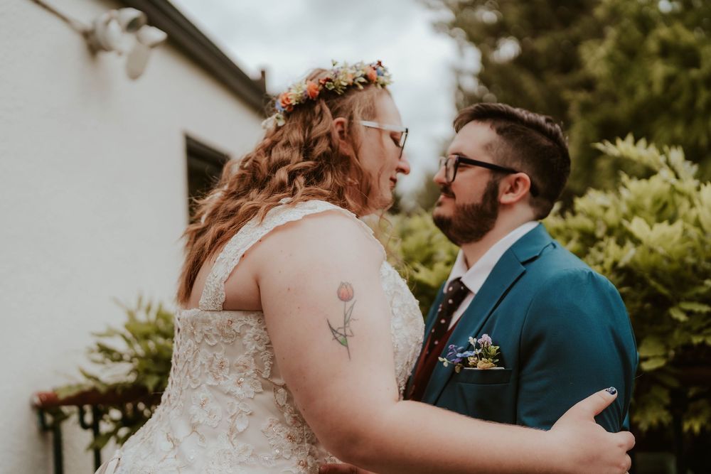 A bride in a white lace gown and floral crown embraces a groom in a teal suit outdoors.