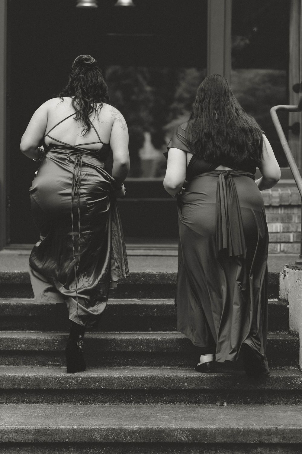 Two bridesmaids in elegant satin gowns walking up steps from behind, black and white wedding photo.
