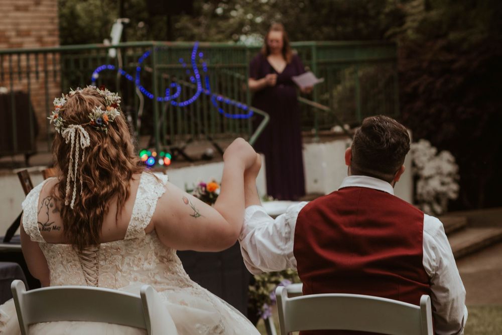 A bride and groom hold hands while seated facing their officiant during an outdoor wedding ceremony.