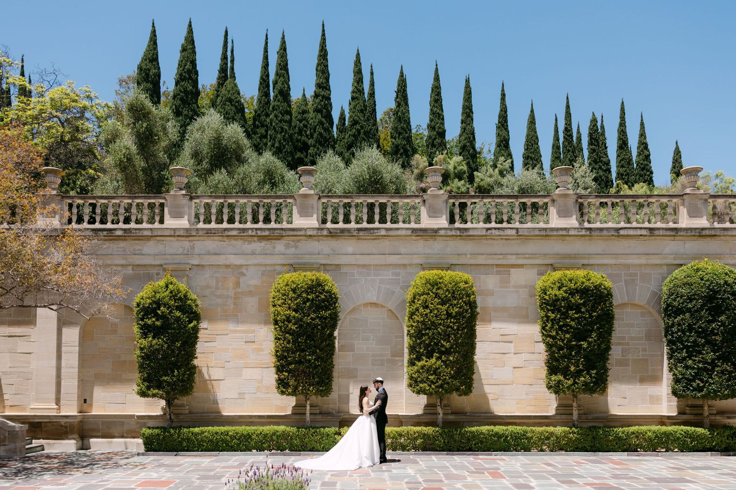 A Mediterranean garden walkway with a row of tall cypress trees and stone walls in an upscale estate setting.