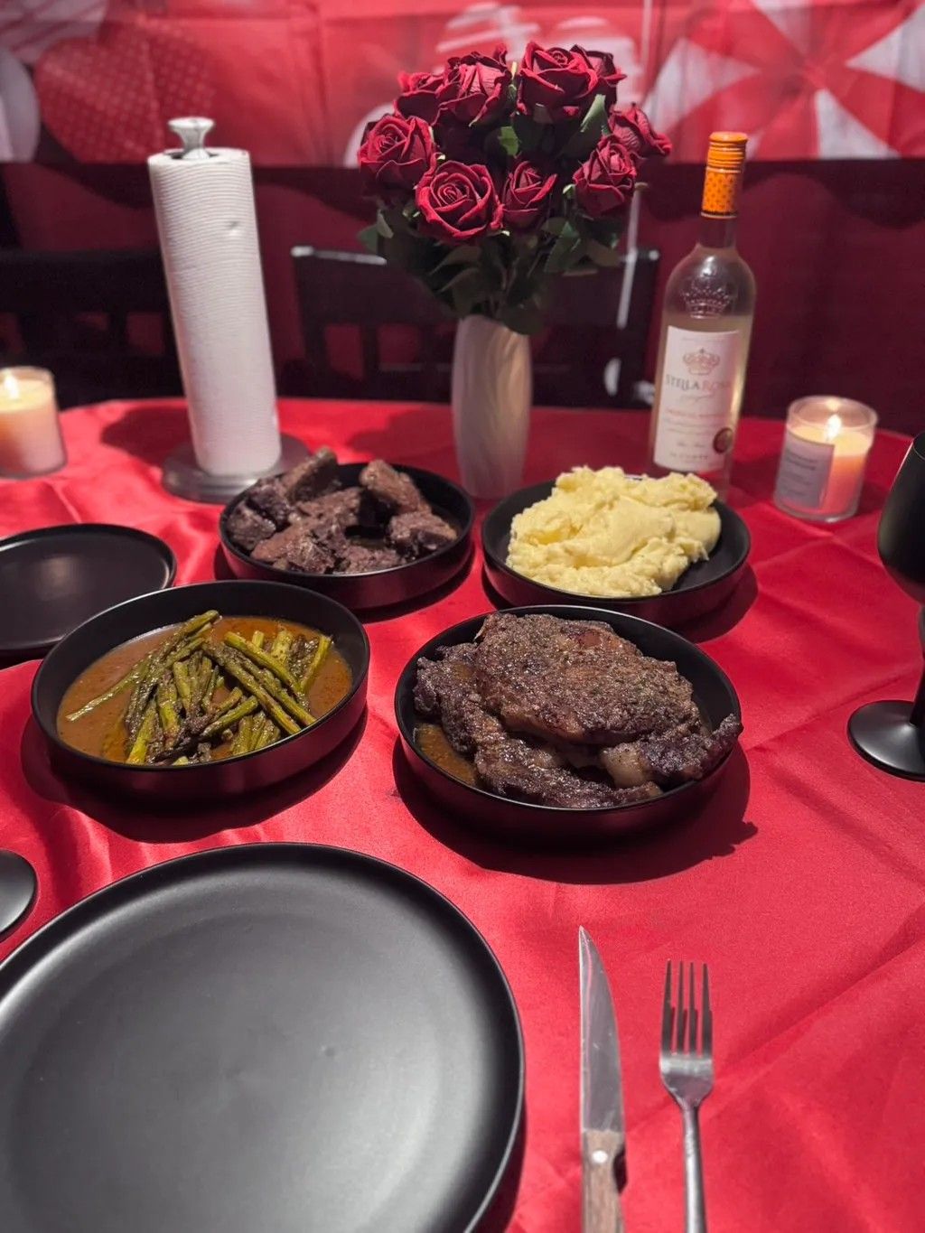 Romantic dinner setting with red roses, wine bottle, candles and various dishes served on black plates over a red tablecloth.