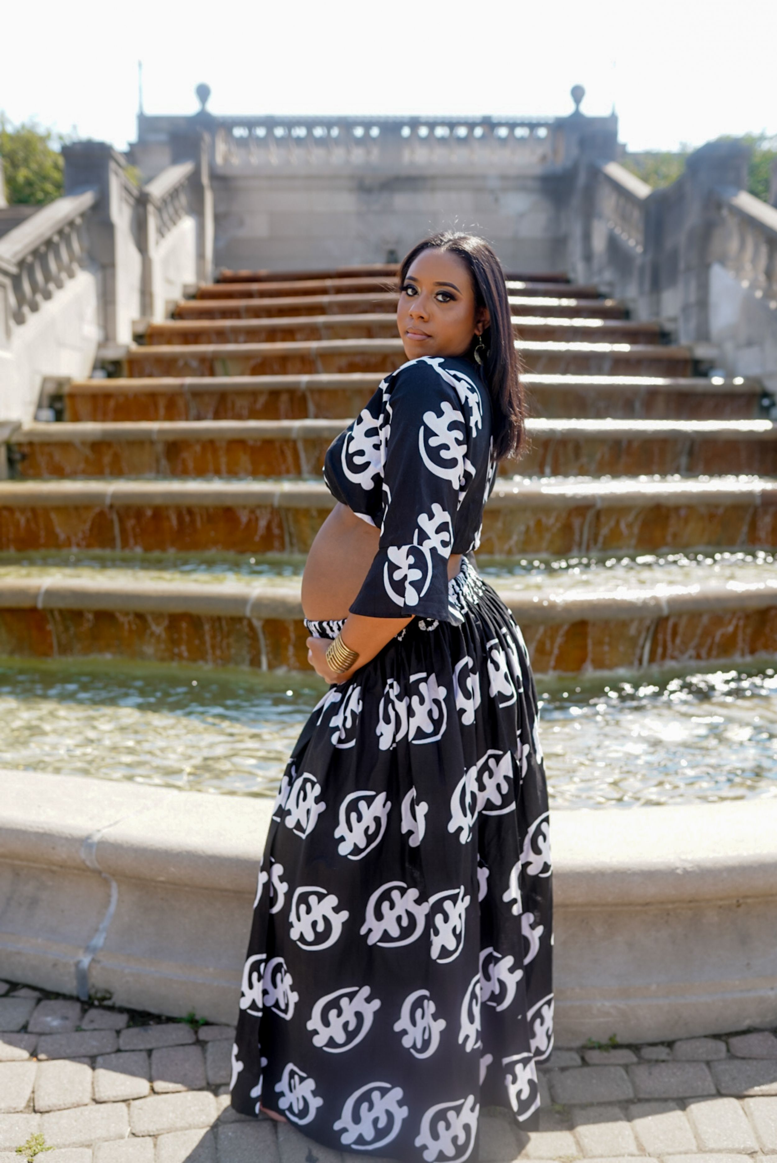 Person in a black and white patterned dress posing by ornate stone fountain steps.