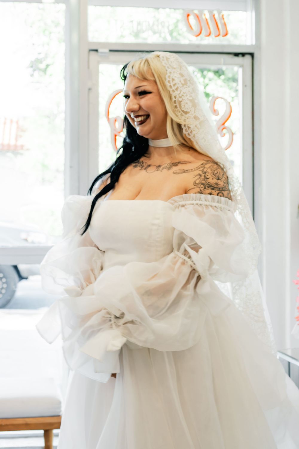 Joyful bride in white off-shoulder tulle gown and lace veil laughing, showcasing tattoos on her shoulder.