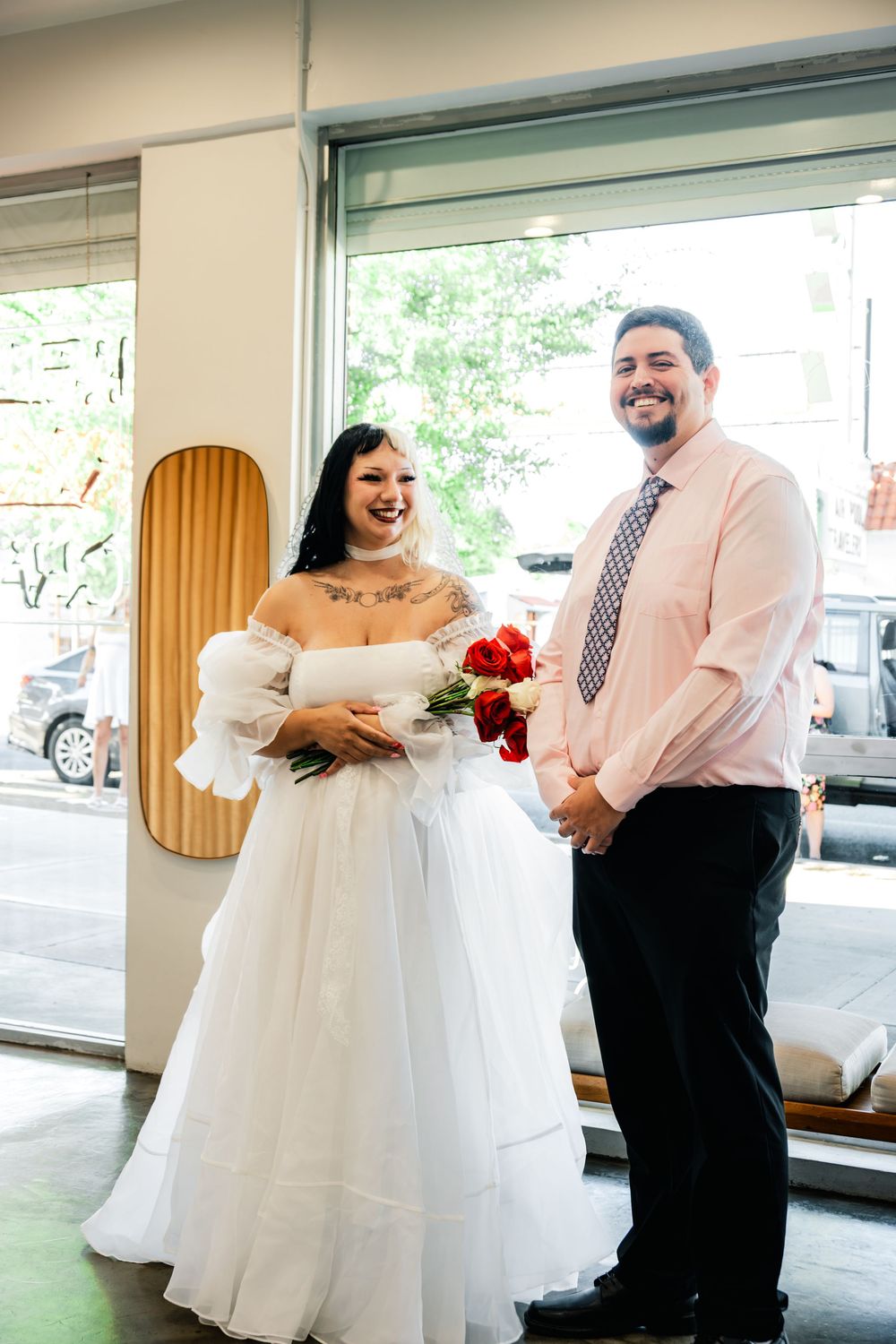 Happy couple posing together on wedding day, bride in white gown holding red roses bouquet.