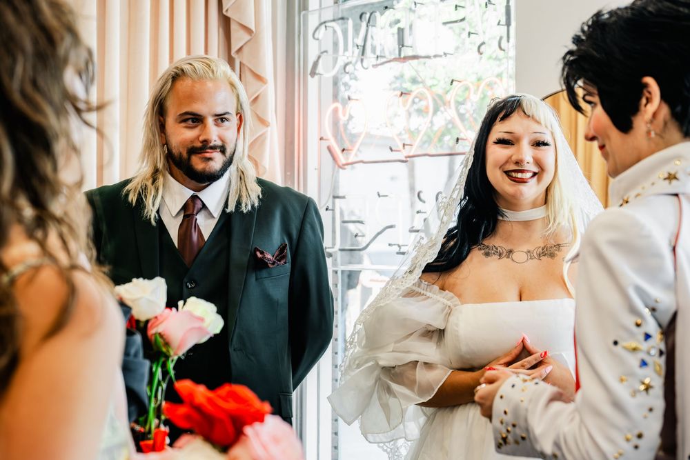 Bride and groom laughing during intimate wedding ceremony with guests holding colorful flowers.