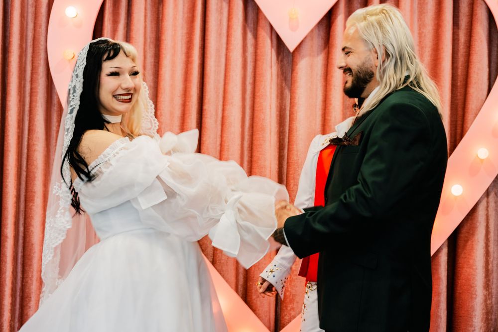 Bride in white dress laughing joyfully with groom before illuminated heart backdrop at wedding.