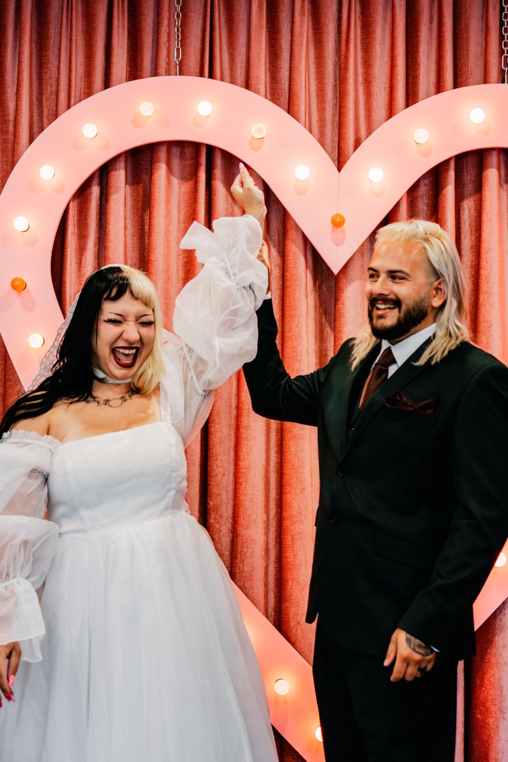Newlyweds celebrate joyfully raising hands before glowing marquee heart backdrop at Las Vegas wedding venue.