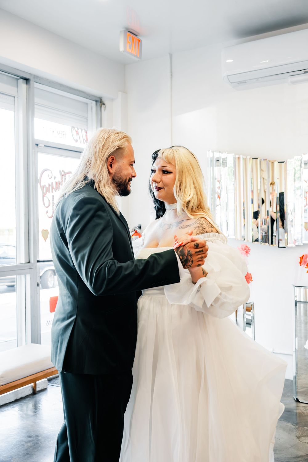 Groom with long blonde hair embraces smiling blonde bride in white gown inside bright wedding venue.
