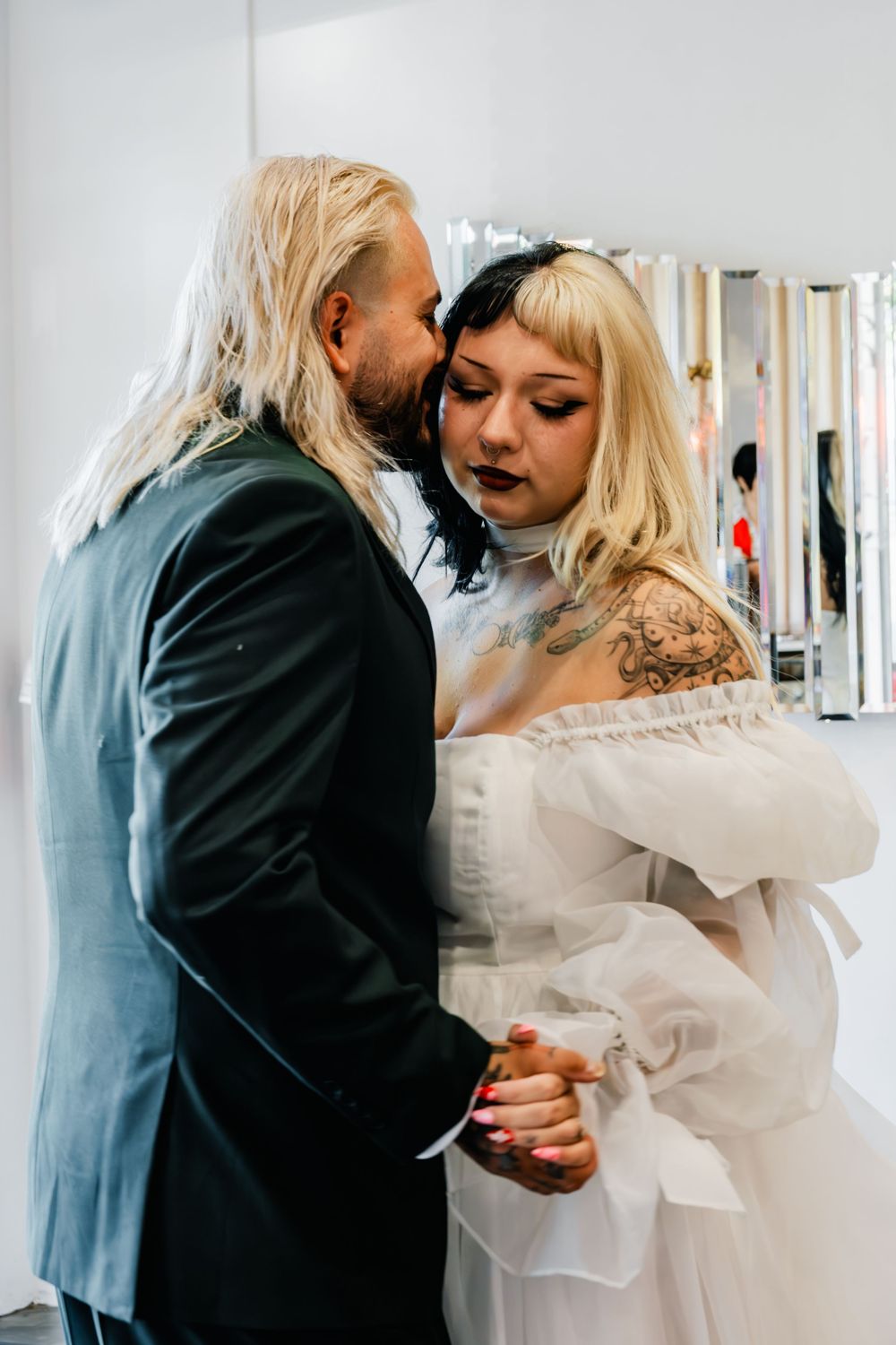 Groom whispers to tattooed blonde bride in white off-shoulder wedding dress during first dance.