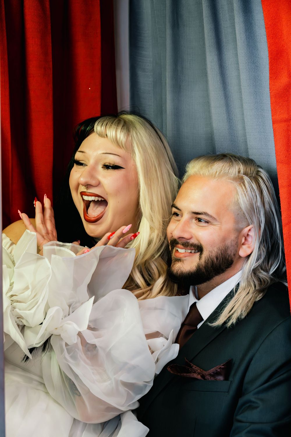 Joyful bride laughing with tongue out beside smiling groom in red-curtained wedding photo booth.