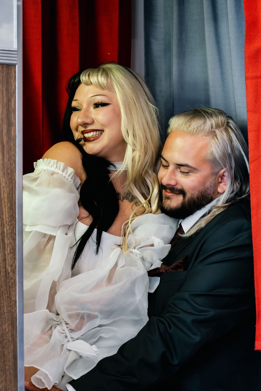 Smiling bride in white ruffled dress embraced by groom in dark suit inside a photo booth with red curtains.