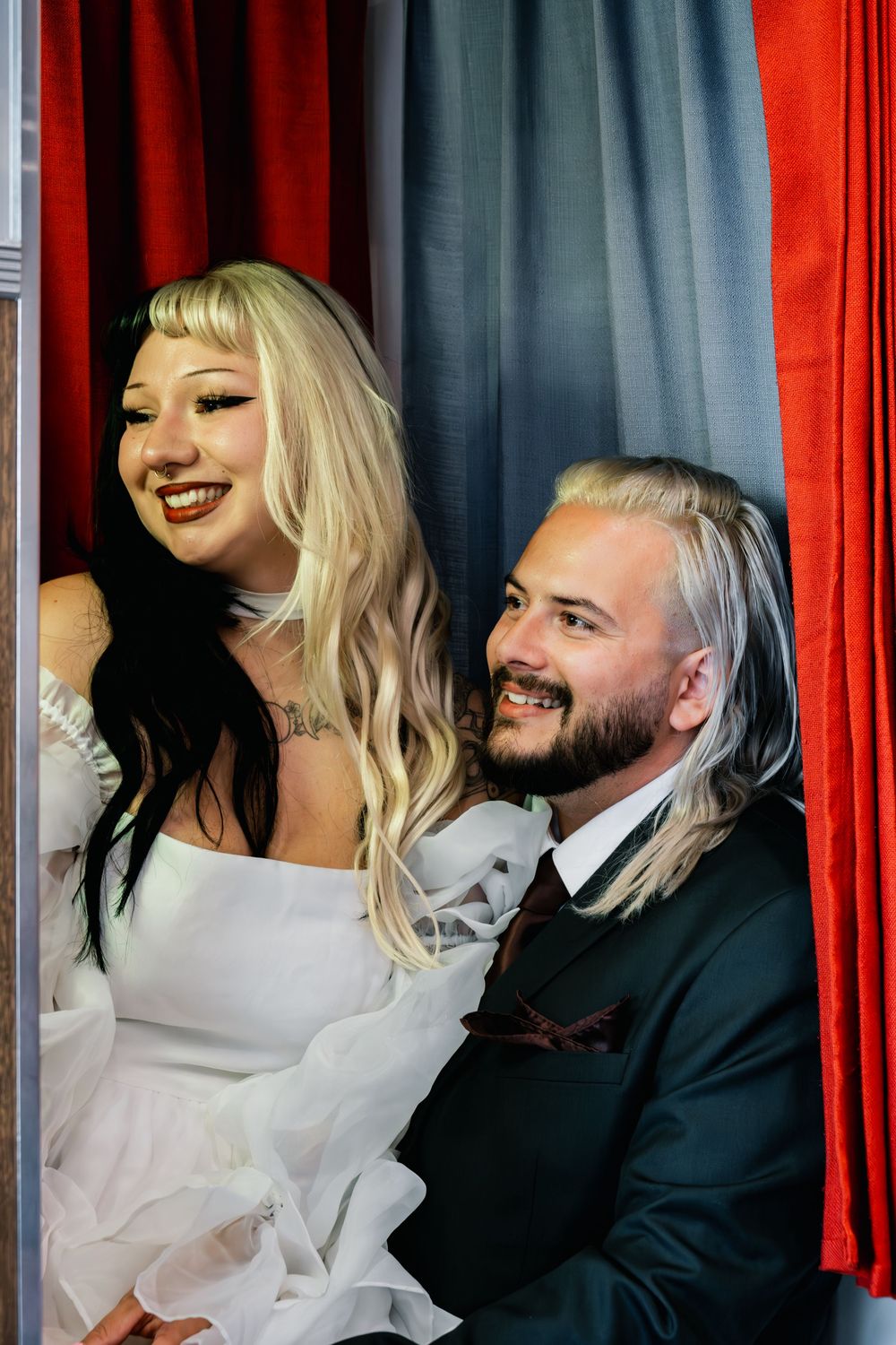 Laughing bride in white dress and smiling groom in dark suit sitting together in a red-curtained photo booth.