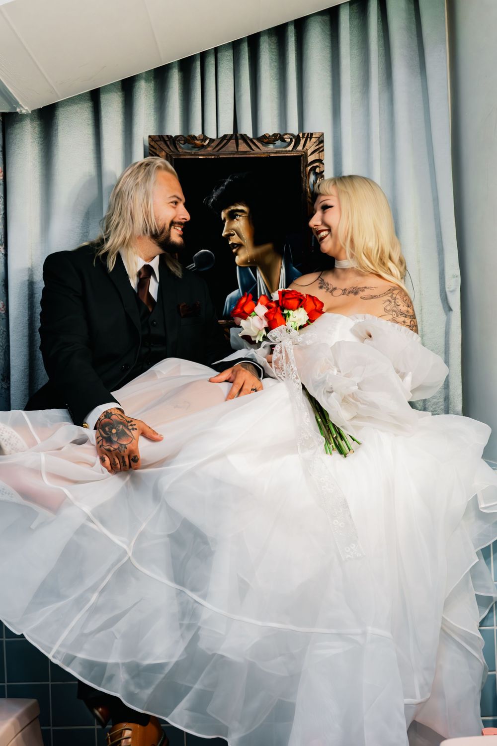 Smiling bride and groom share a joyful moment seated together in front of an Elvis Presley portrait.