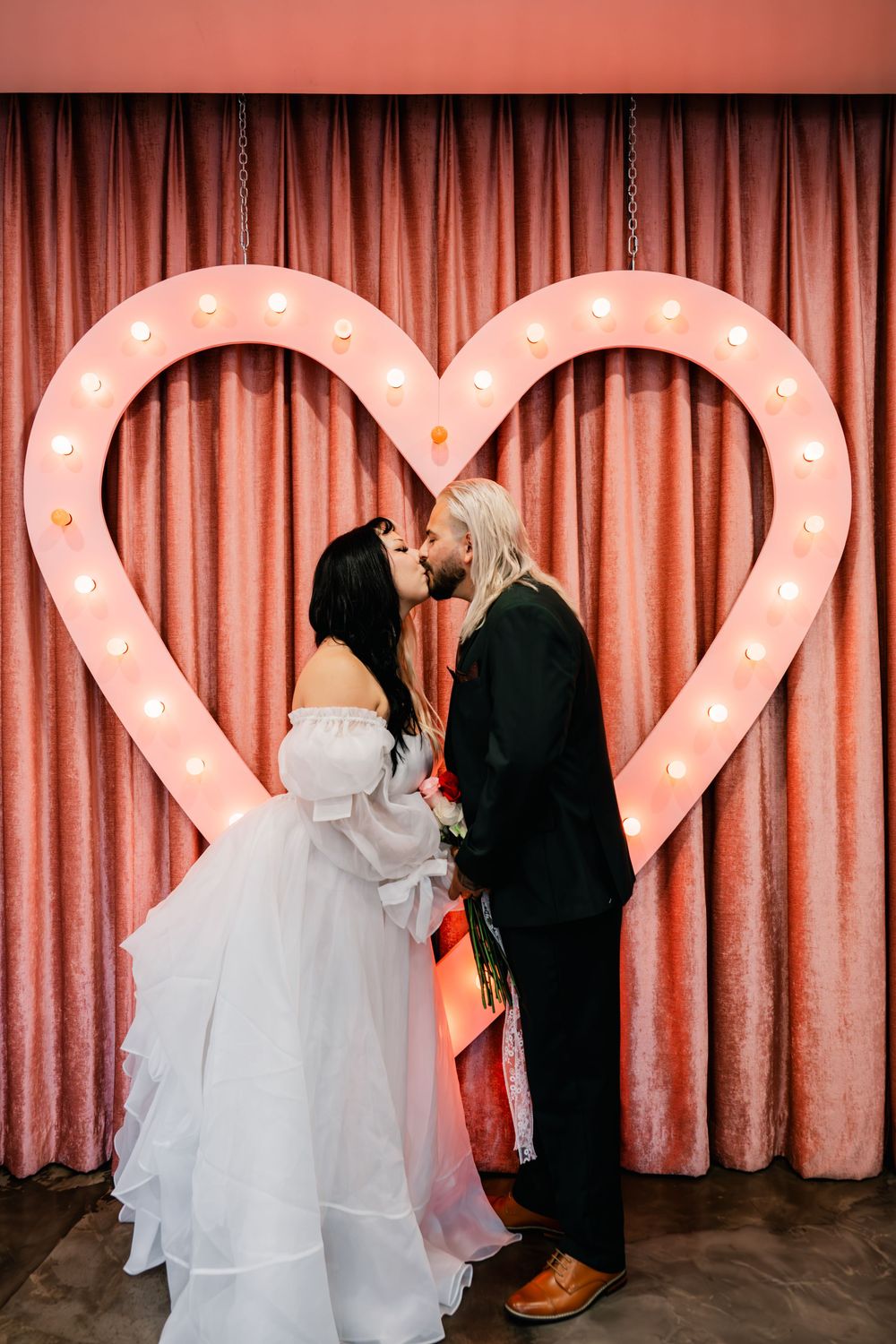Bride and groom sharing first kiss under glowing heart marquee sign with pink velvet curtain background.
