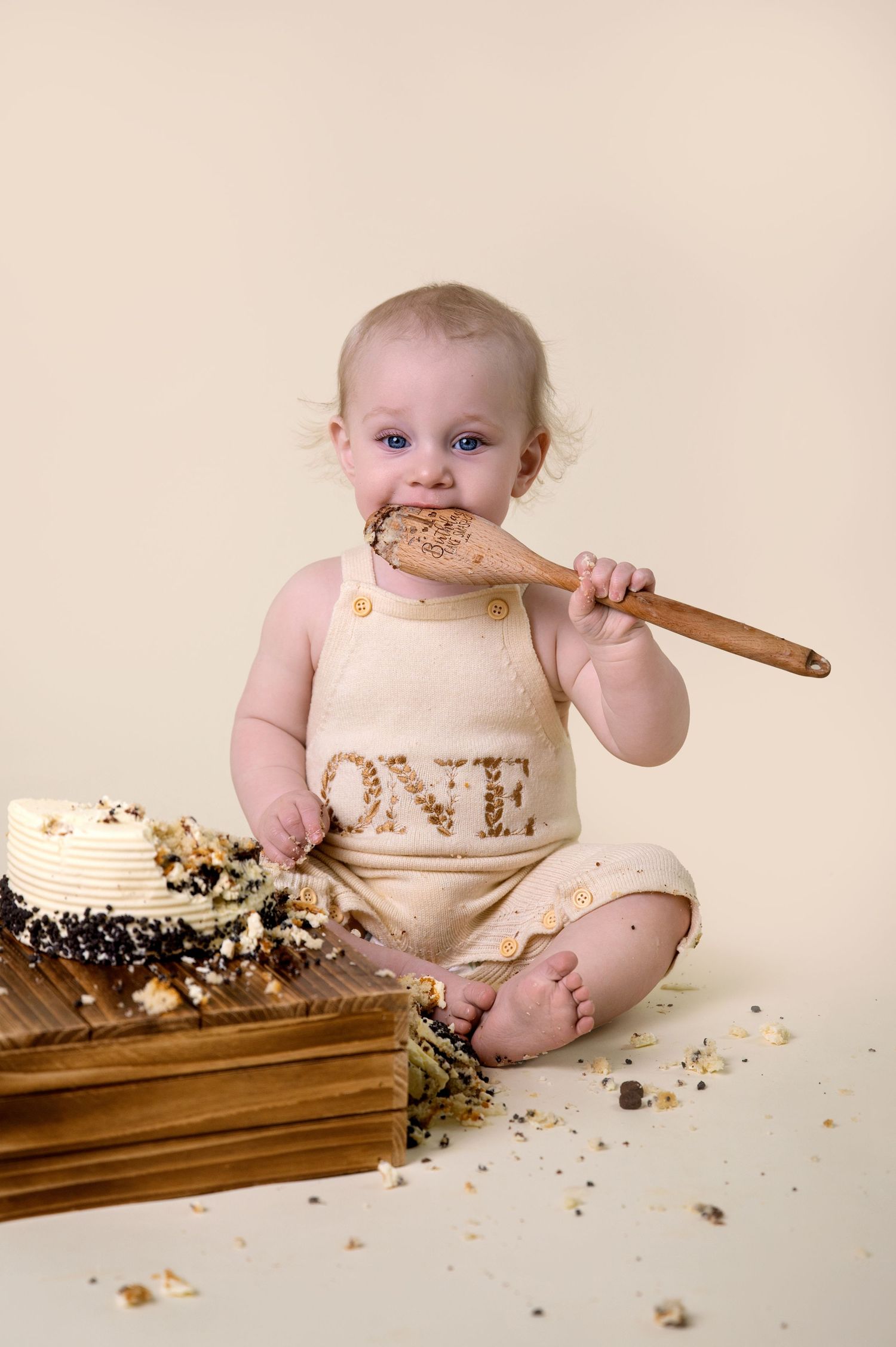 Baby wearing tan colored romper crawls and smiles on cream colored backdrop with stuffed toys nearby.